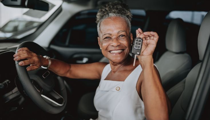 An older woman sitting in a new car, holding up the keys.